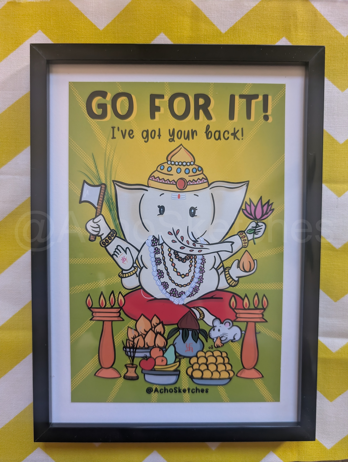 A framed Ganesha art print with the text 'GO FOR IT! I've goi your back!' featuring an illustration of Ganesha on a yellow background, enclosed in a black frame and set against a yellow and white chevron backdrop.